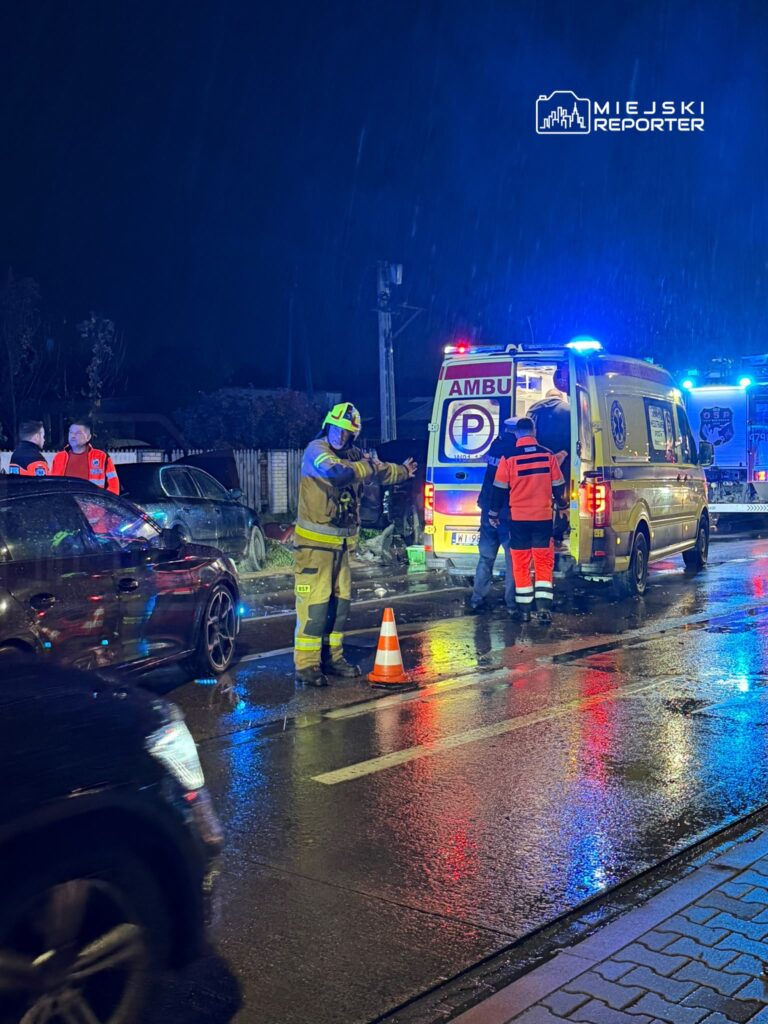 Strażak i ratownicy przy ambulansie na mokrej, oświetlonej sygnałami drodze nocą.