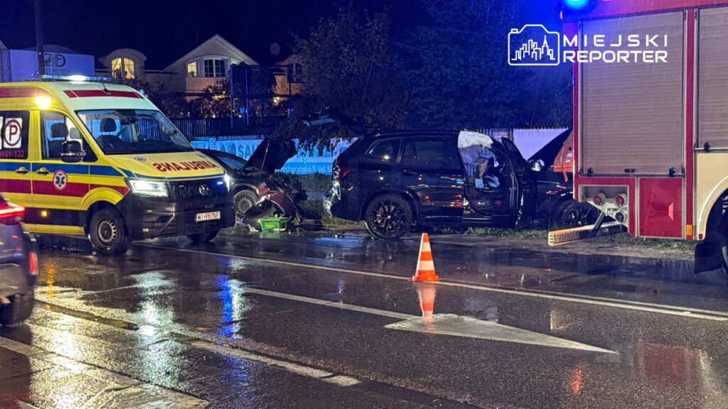 Uszkodzony czarny samochód i karetka przy wozie strażackim na mokrej jezdni, zabezpieczonej pachołkiem drogowym nocą.