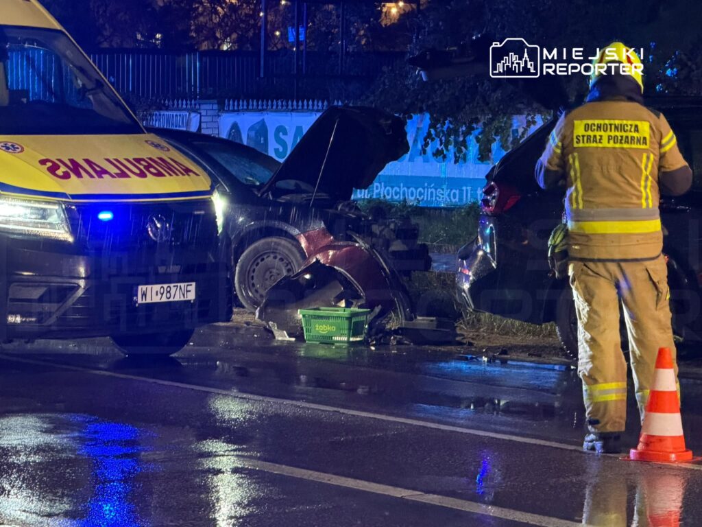 Strażak ochotniczej straży pożarnej stoi przy rozbitym samochodzie i karetce na mokrej ulicy nocą.