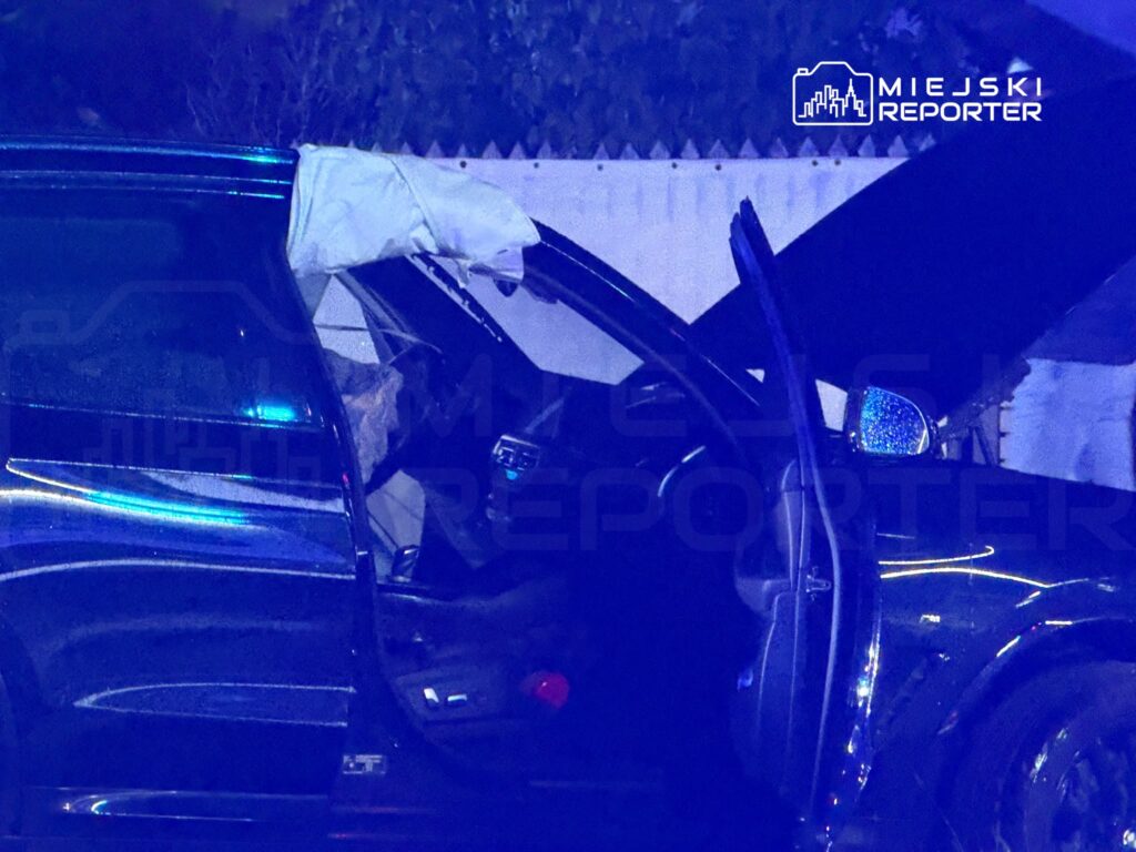 Ciemny samochód z otwartymi drzwiami i uszkodzoną przednią szybą stoi nocą przy ogrodzeniu.
