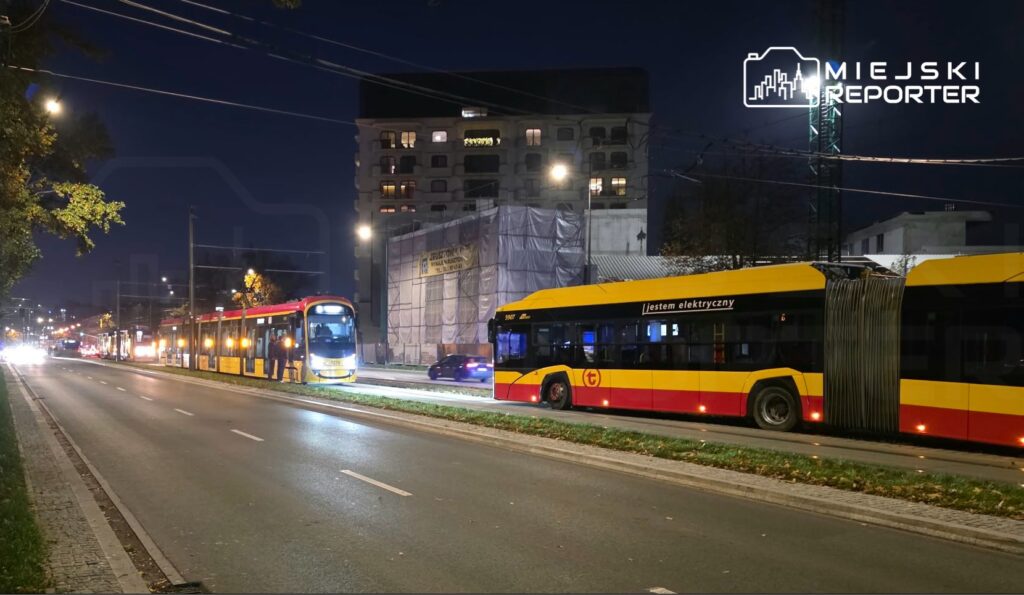Żółto-czerwony autobus elektryczny i tramwaj stoją na nocnej ulicy przy torowisku.