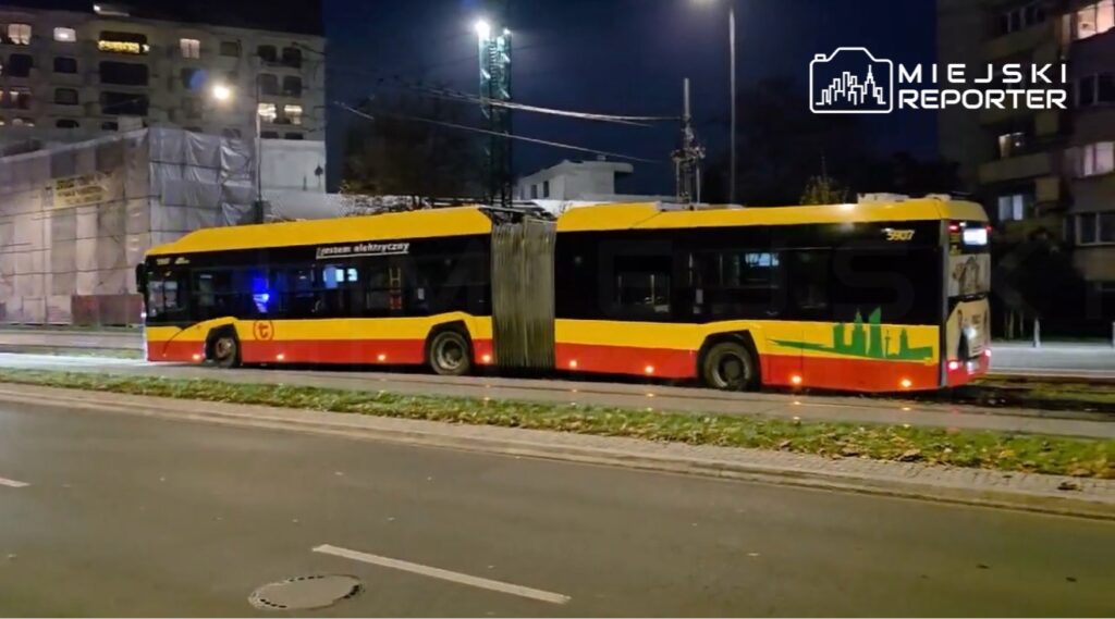 Żółto-czerwony przegubowy autobus miejski stoi nocą na ulicy, obok oświetlonych budynków.