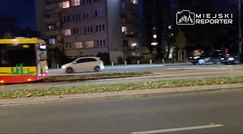 Kolorowy tramwaj i samochody jadą nocą ulicą, mijając blok mieszkalny.