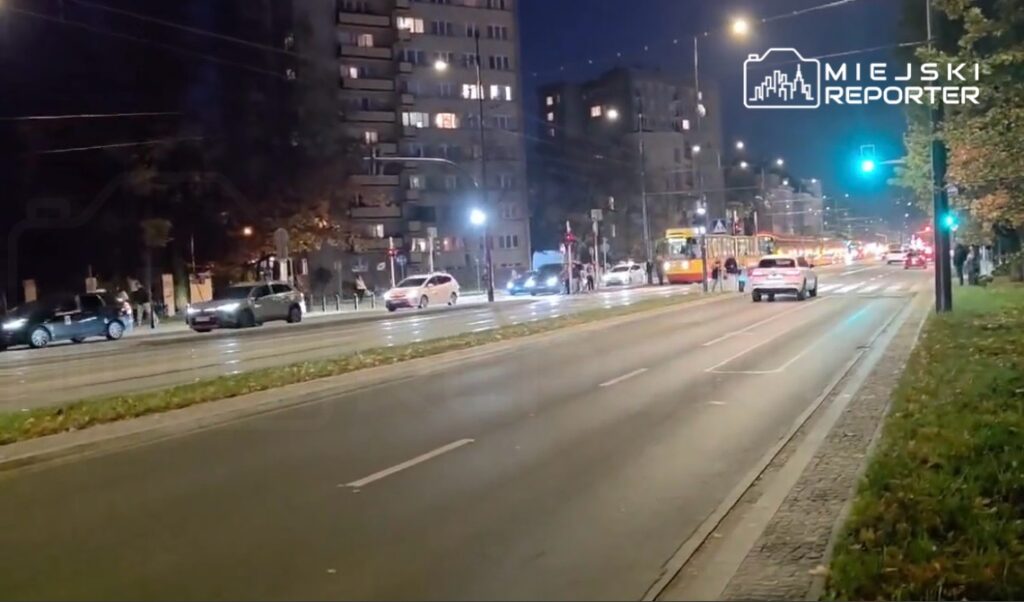 Samochody i tramwaj jadą nocą miejską ulicą, mijając bloki mieszkalne i zielone światło sygnalizacji.