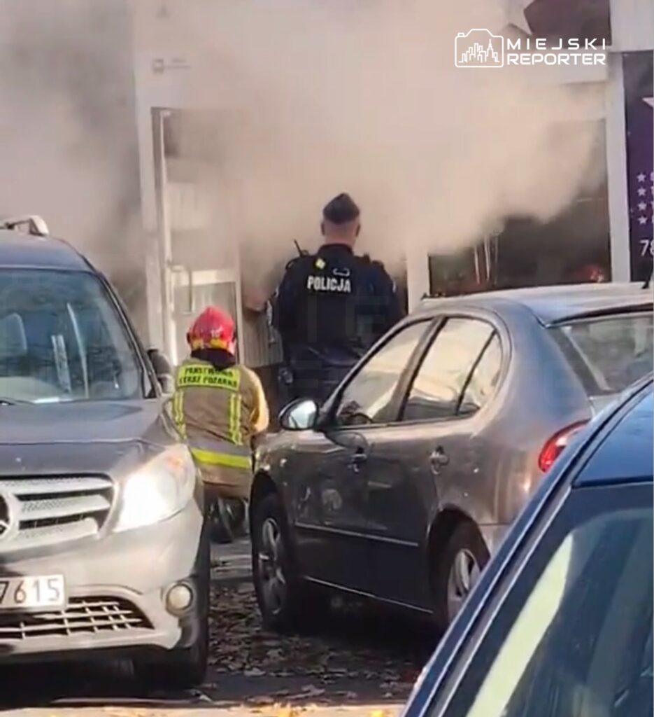 Policjant i strażak stoją przed zadymionym wejściem do budynku, między zaparkowanymi samochodami.