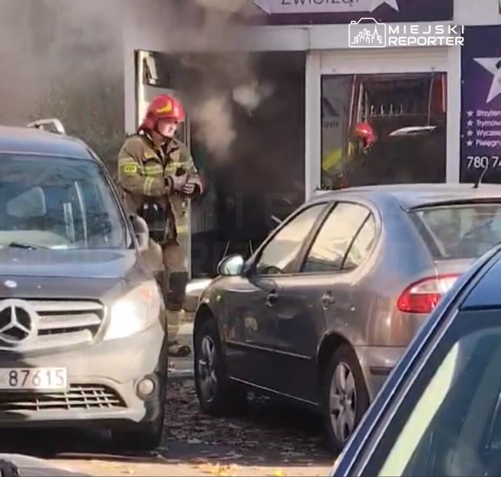 Strażak w czerwonym hełmie wychodzi z zadymionego lokalu między zaparkowanymi samochodami.