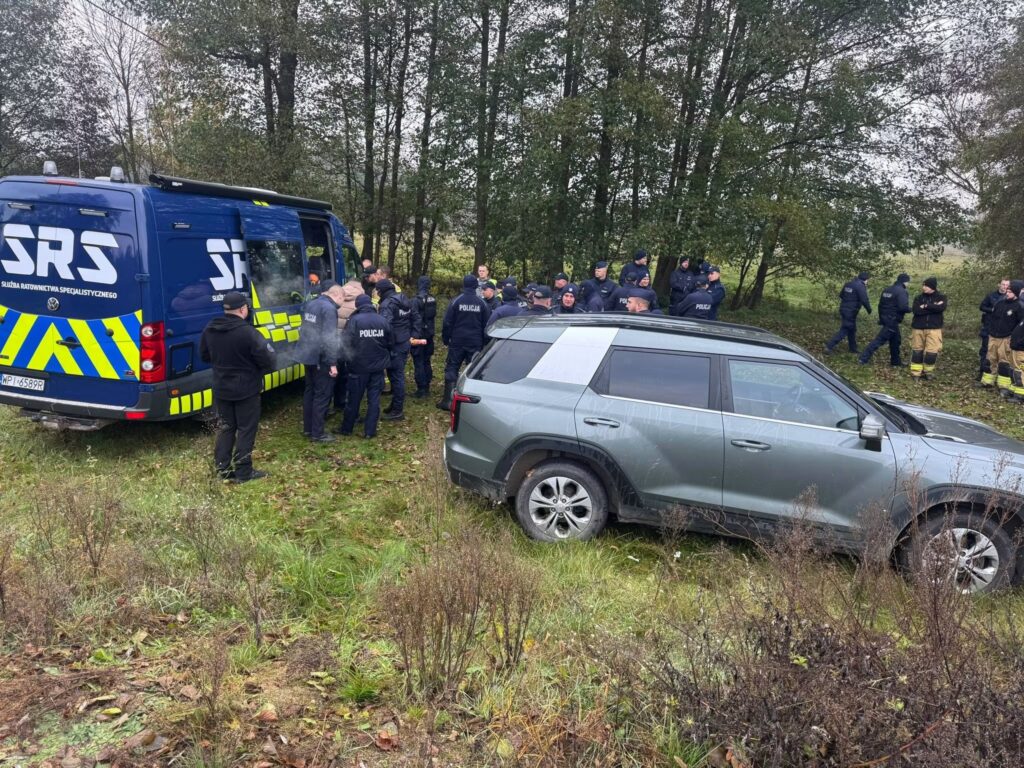 Policjanci i służby ratunkowe stoją przy niebieskim busie SRS i szarym SUV-ie na leśnej drodze.