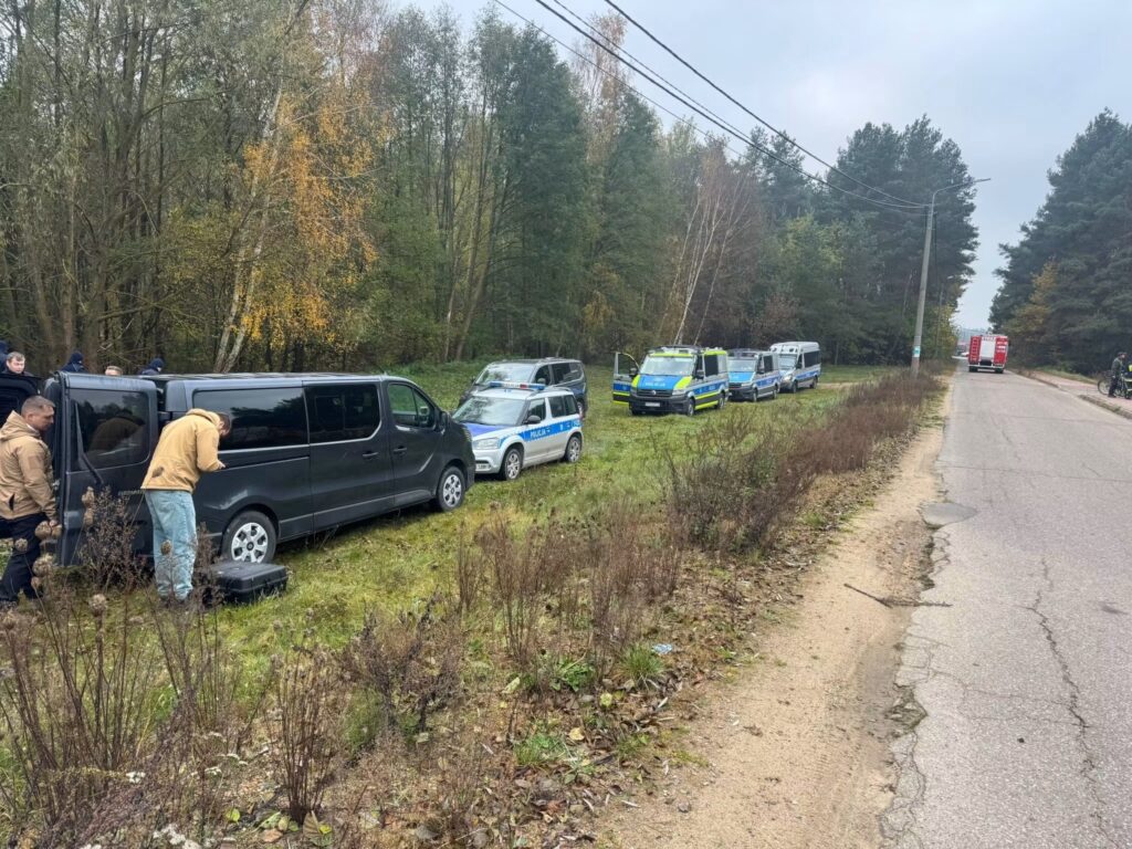 Samochody służb ratunkowych i osobowe stoją na poboczu leśnej drogi, a ludzie gromadzą się przy ciemnym vanie.