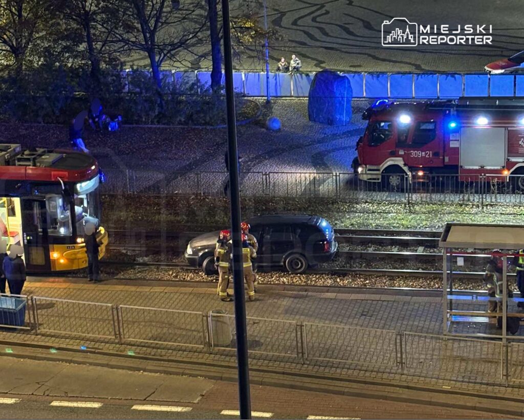 Strażacy zabezpieczają samochód stojący na torowisku, obok tramwaju i wozu strażackiego z włączonymi światłami.