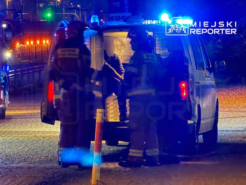 Ratownicy w odblaskowych ubraniach udzielają pomocy osobie w otwartych drzwiach ambulansu, oświetlonego niebieskimi
