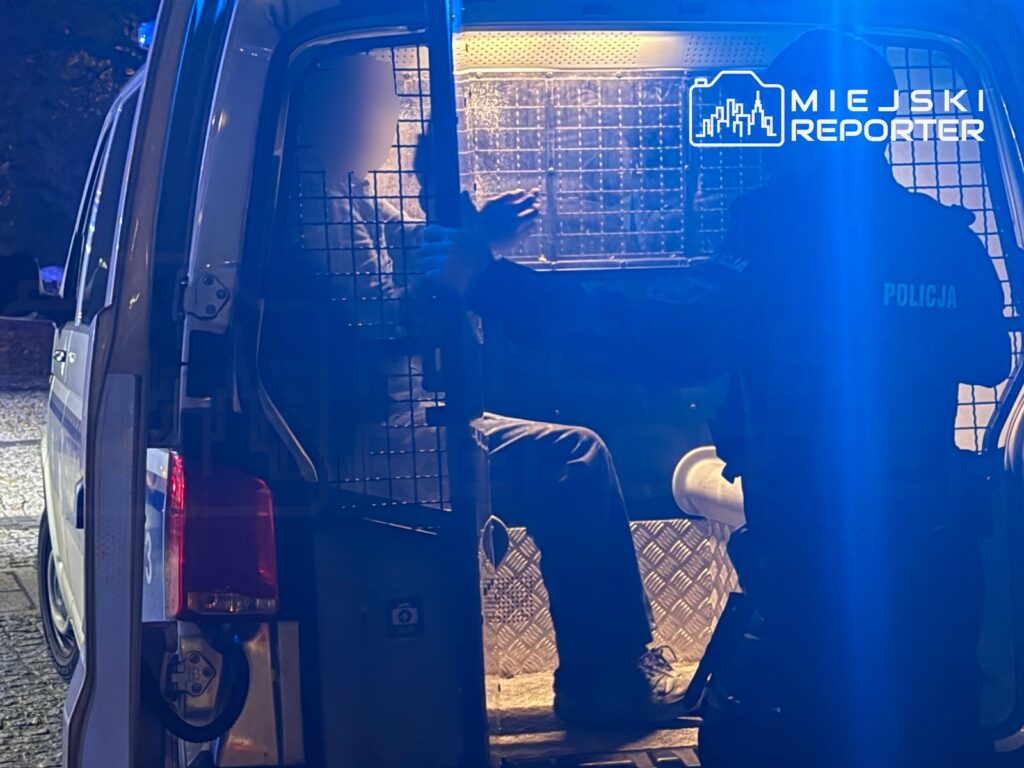 Policjant prowadzi mężczyznę do radiowozu z otwartymi drzwiami, oświetlonego niebieskim światłem.