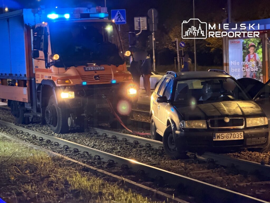 Ciężarówka służb technicznych stoi na torach obok samochodu osobowego, nocą przy przejeździe kolejowym.