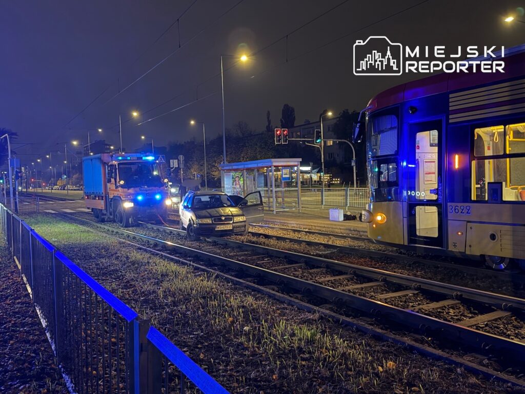 Samochód osobowy stoi na torowisku między tramwajem a pojazdem służb ratunkowych, nocą przy przystanku tramwajowym.