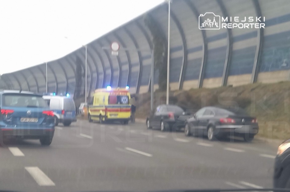 Karetka pogotowia z włączonymi światłami stoi na jezdni przy zaparkowanych samochodach i wysokim ekranie akustycznym.