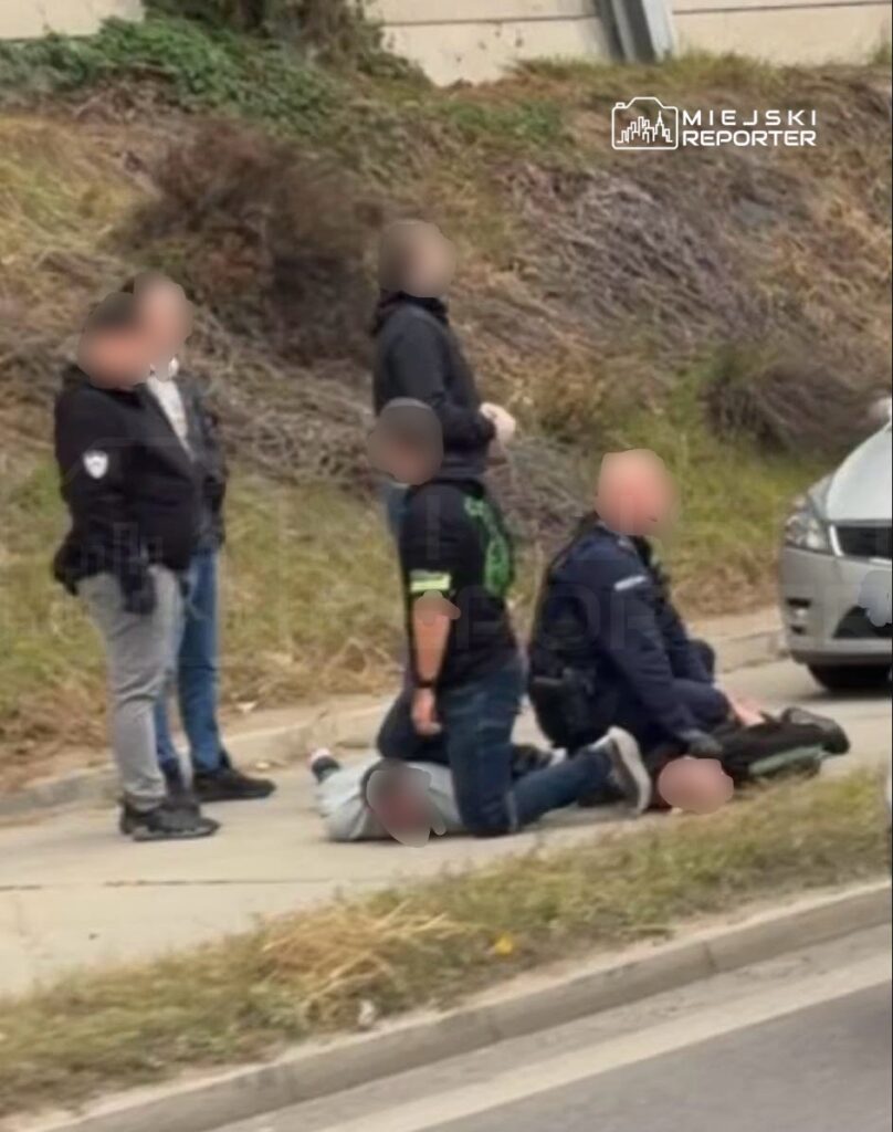 Trzech mężczyzn stoi i klęczy przy osobie leżącej na chodniku, obok zaparkowanego samochodu.