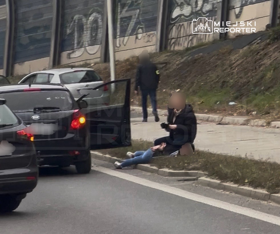 Osoba siedzi na chodniku przy leżącej osobie, obok zatrzymanych samochodów i otwartych drzwi auta.