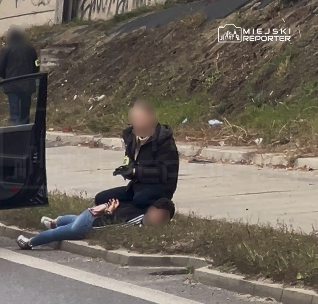 Mężczyzna klęczy obok leżącej osoby przy krawężniku, a drugi stoi przy otwartych drzwiach pojazdu.