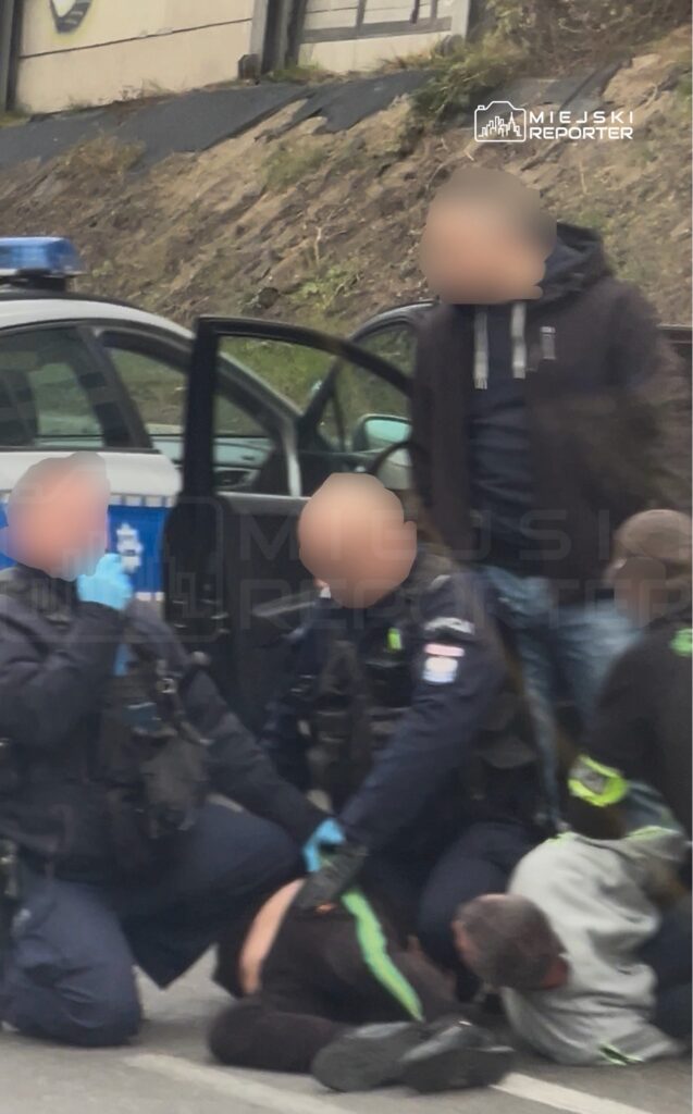 Policjanci obezwładniają mężczyznę na chodniku obok radiowozu, a drugi funkcjonariusz stoi przy otwartych drzwiach auta.