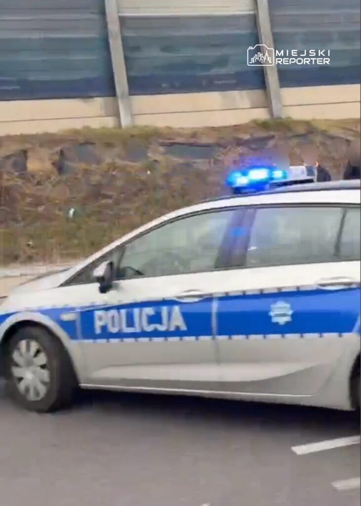 Radiowóz z napisem „POLICJA” i włączonymi niebieskimi sygnałami świetlnymi na drodze przy ekranie akustycznym.