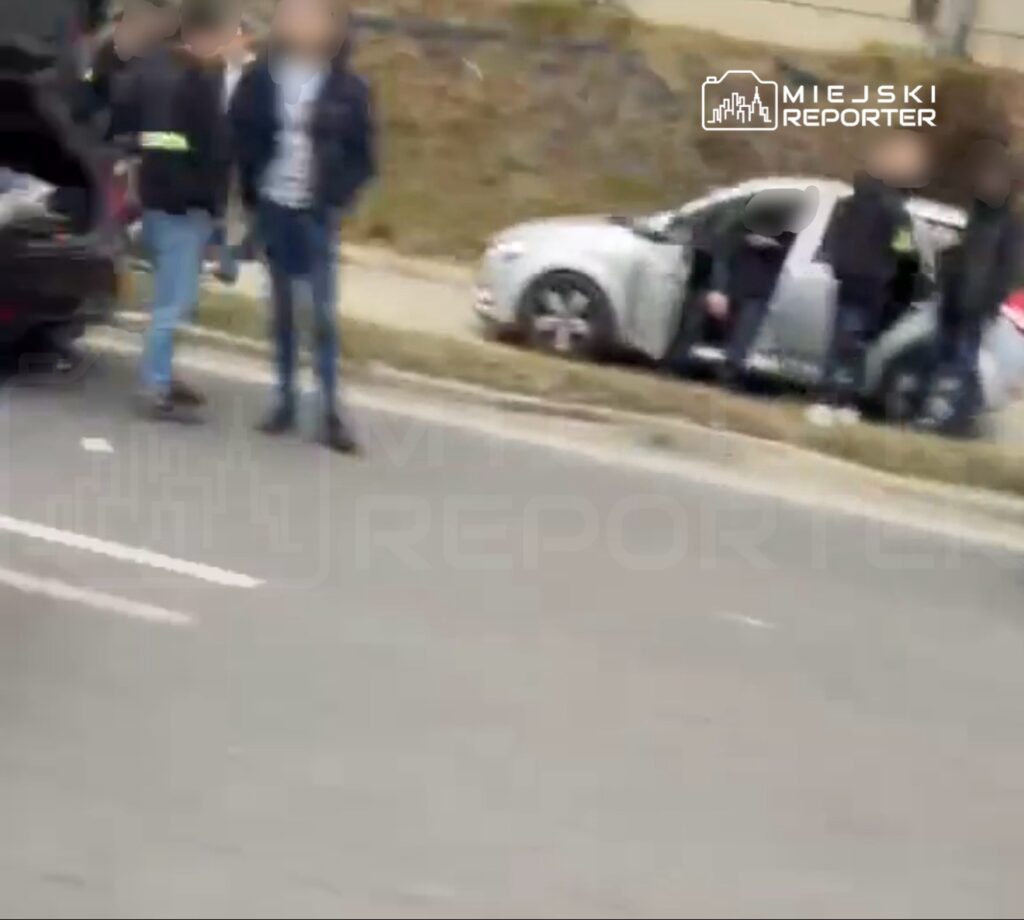 Osoby stoją przy srebrnym samochodzie z otwartymi drzwiami na poboczu drogi.