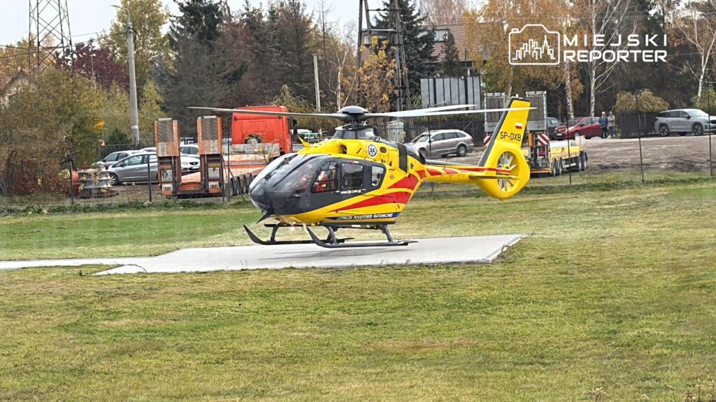 Żółto-czerwony śmigłowiec ratunkowy stoi na betonowym lądowisku na trawiastym terenie, za nim widać parking i drzewa.