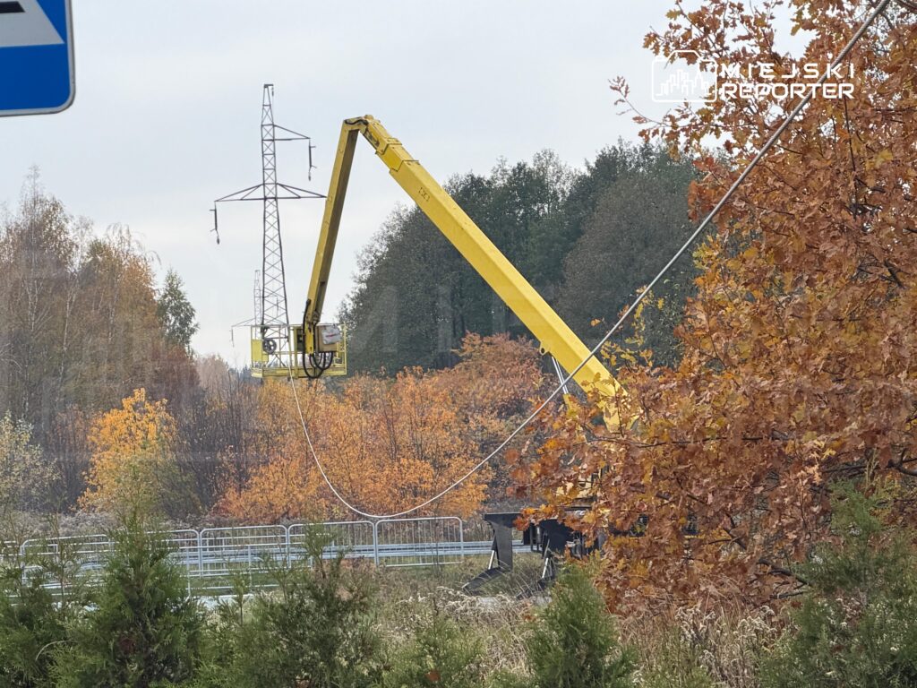 Żółty podnośnik koszowy przy słupie energetycznym, otoczony jesiennymi drzewami i barierkami.