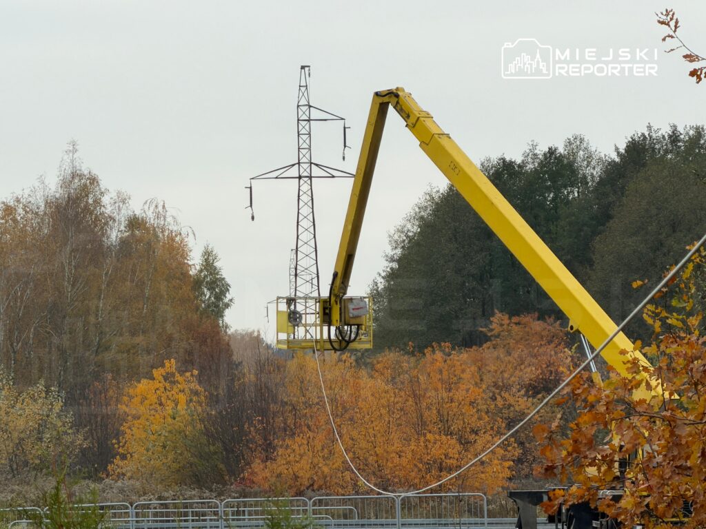Żółty podnośnik z koszem unosi pracownika przy stalowym słupie energetycznym pośród jesiennych drzew.
