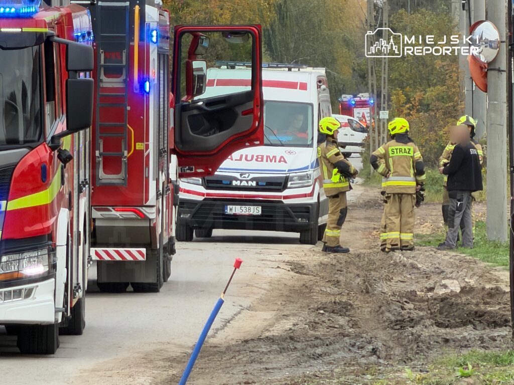 Strażacy w odblaskowych mundurach rozmawiają przy drodze obok wozu strażackiego i karetki pogotowia.