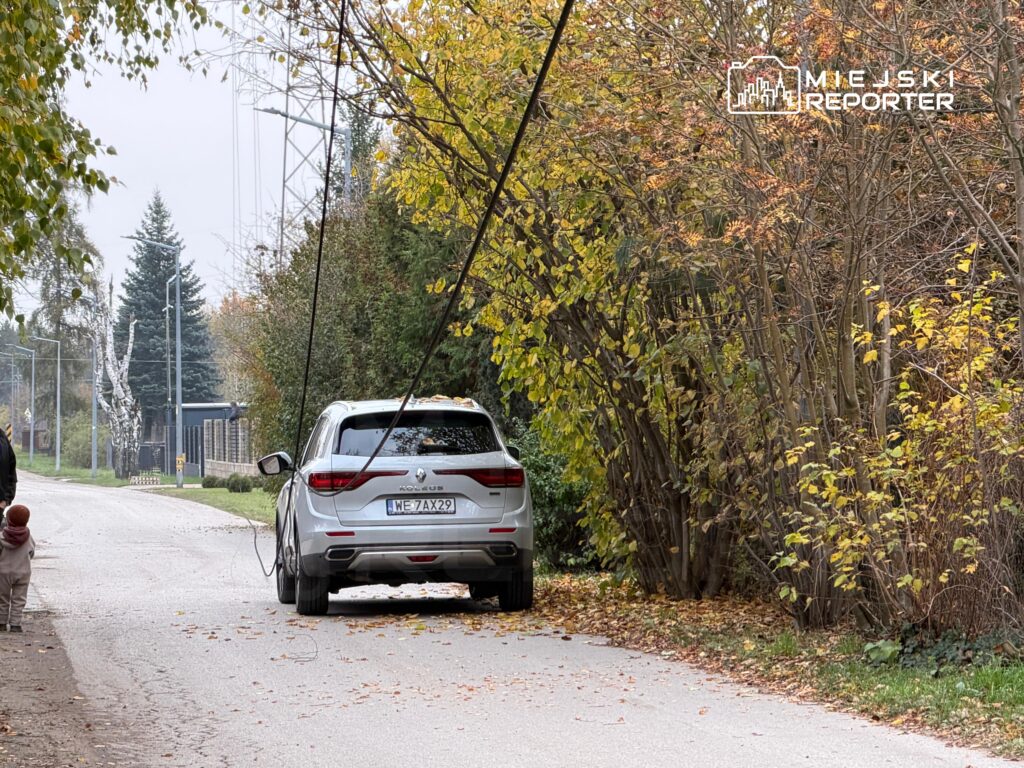 Biały SUV stoi na wąskiej drodze przy zaroślach i drzewach z jesiennymi liśćmi.