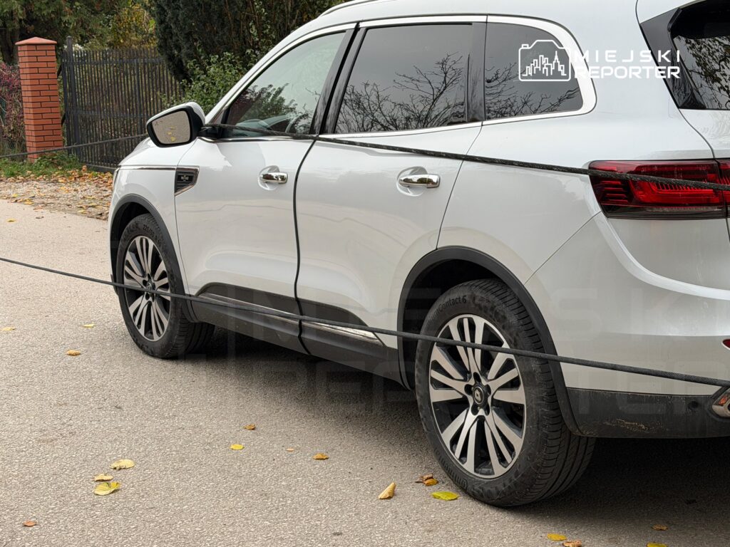 Biały SUV zaparkowany przy ogrodzeniu, widoczny z boku na tle jesiennej ulicy.