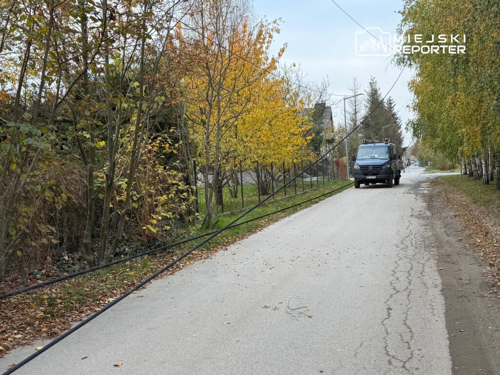 Czarna ciężarówka stoi na wąskiej, asfaltowej drodze wśród jesiennych drzew; przez jezdnię biegnie zwisający przewód.