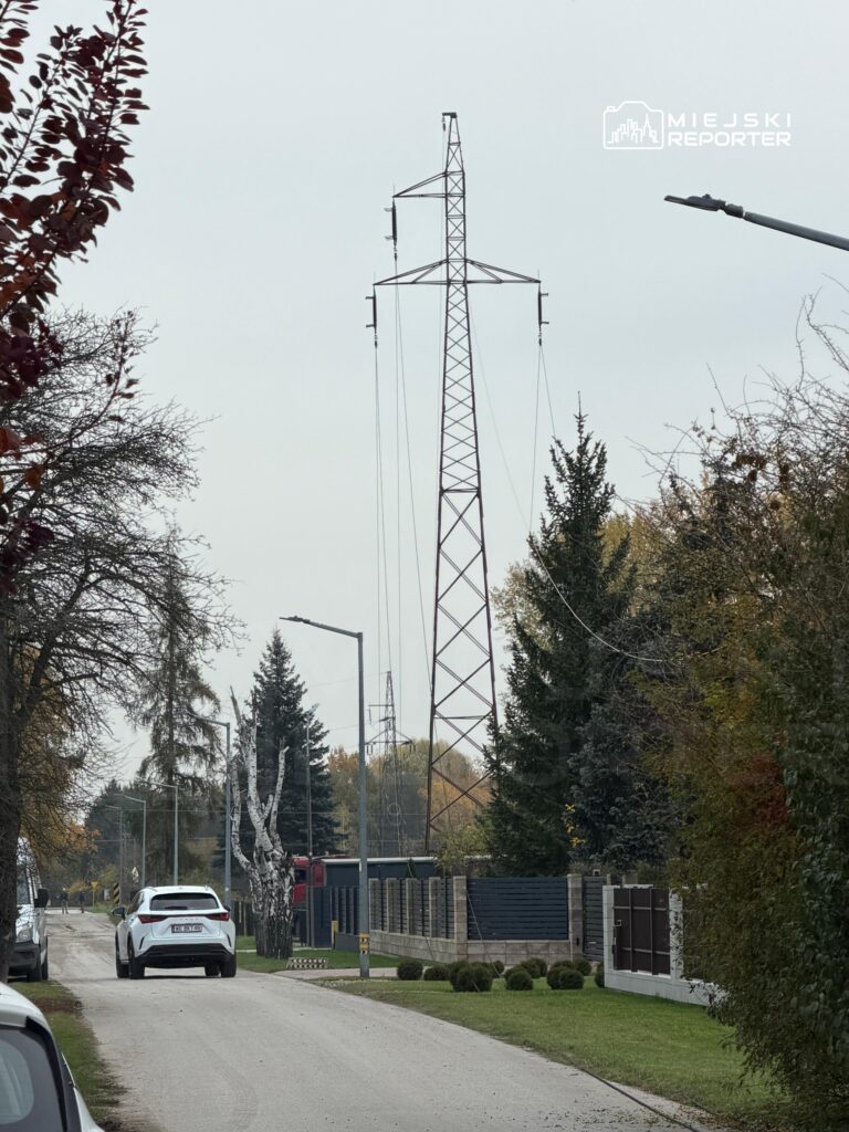 Wysoki metalowy słup energetyczny przy wąskiej ulicy z zaparkowanymi samochodami i domami otoczonymi drzewami.