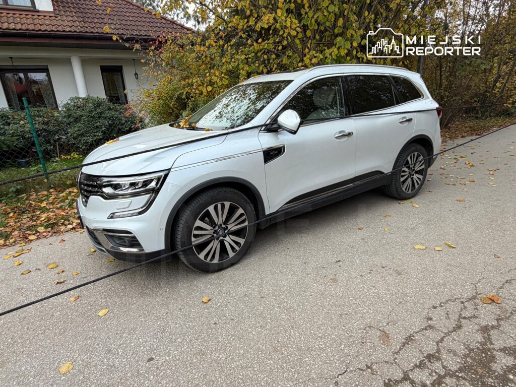 Biały SUV zaparkowany przy jezdni przed domem, jesienne liście leżą na drodze i gałęziach drzewa.