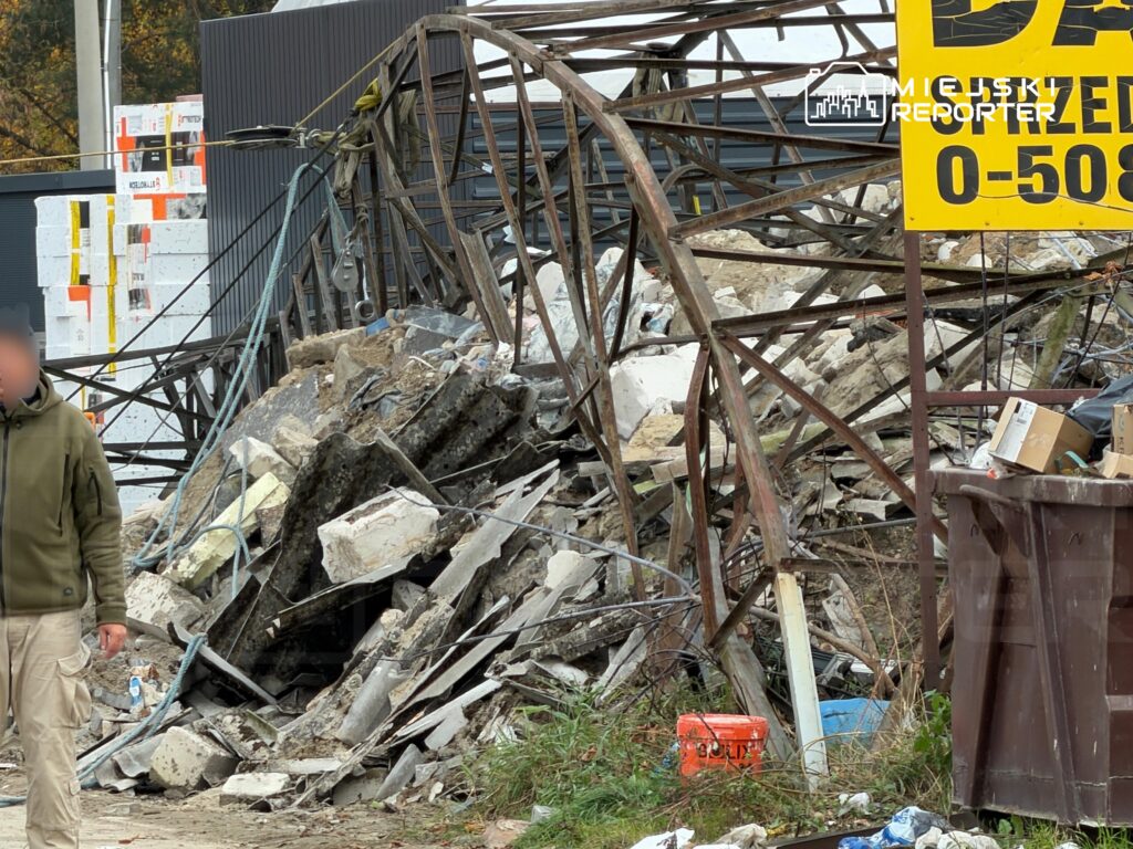 Metalowa konstrukcja zawalona na stertę gruzu i betonowych bloków obok placu budowy; z lewej stoi mężczyzna.