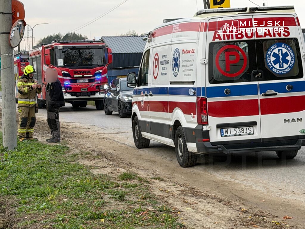 Strażak rozmawia z osobą przy drodze, obok stoją karetka pogotowia i czerwony wóz strażacki.