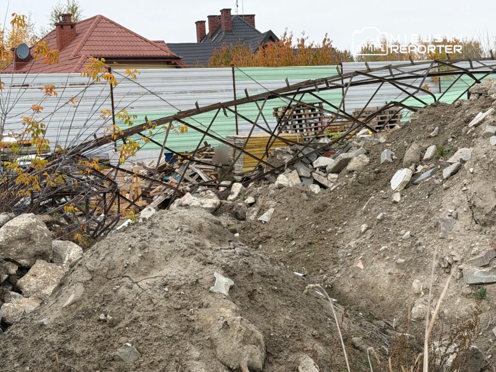 Zawalona metalowa konstrukcja ogrodzenia leży na gruzie i zwałach ziemi przy zabudowaniach mieszkalnych.