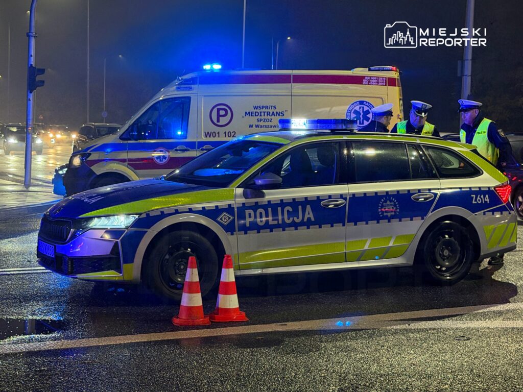 Radiowóz policyjny i karetka z włączonymi światłami stoją na mokrej ulicy, obok funkcjonariusze i pachołki drogowe.