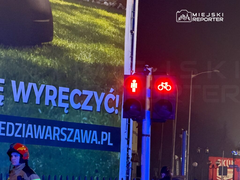 Czerwone sygnalizatory dla pieszych i rowerzystów świecą nocą obok reklamowego billboardu.