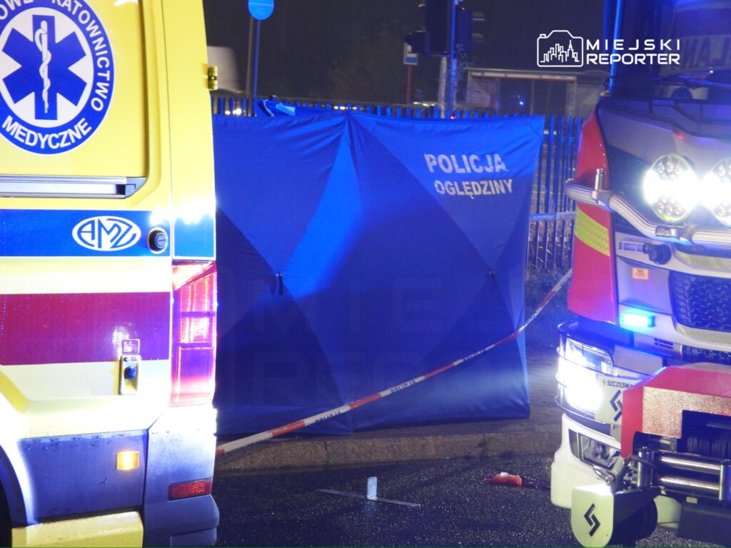Niebieski parawan z napisem „POLICJA OGLĘDZINY” między karetką a wozem strażackim na nocnym miejscu zdarzenia.