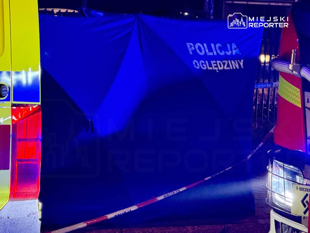 Niebieski parawan z napisem „POLICJA OGLĘDZINY” między radiowozami, odgrodzony taśmą policyjną.
