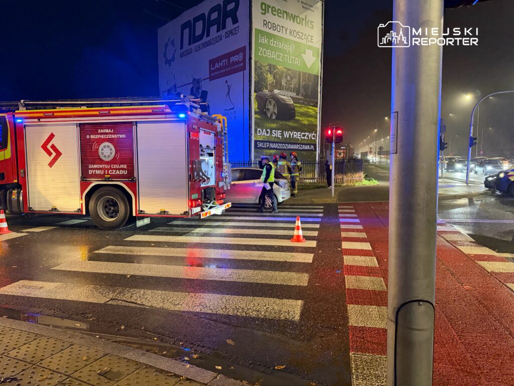 Wóz strażacki z włączonymi światłami stoi na mokrym przejściu dla pieszych, obok pracują ratownicy i ustawiono pachołek.