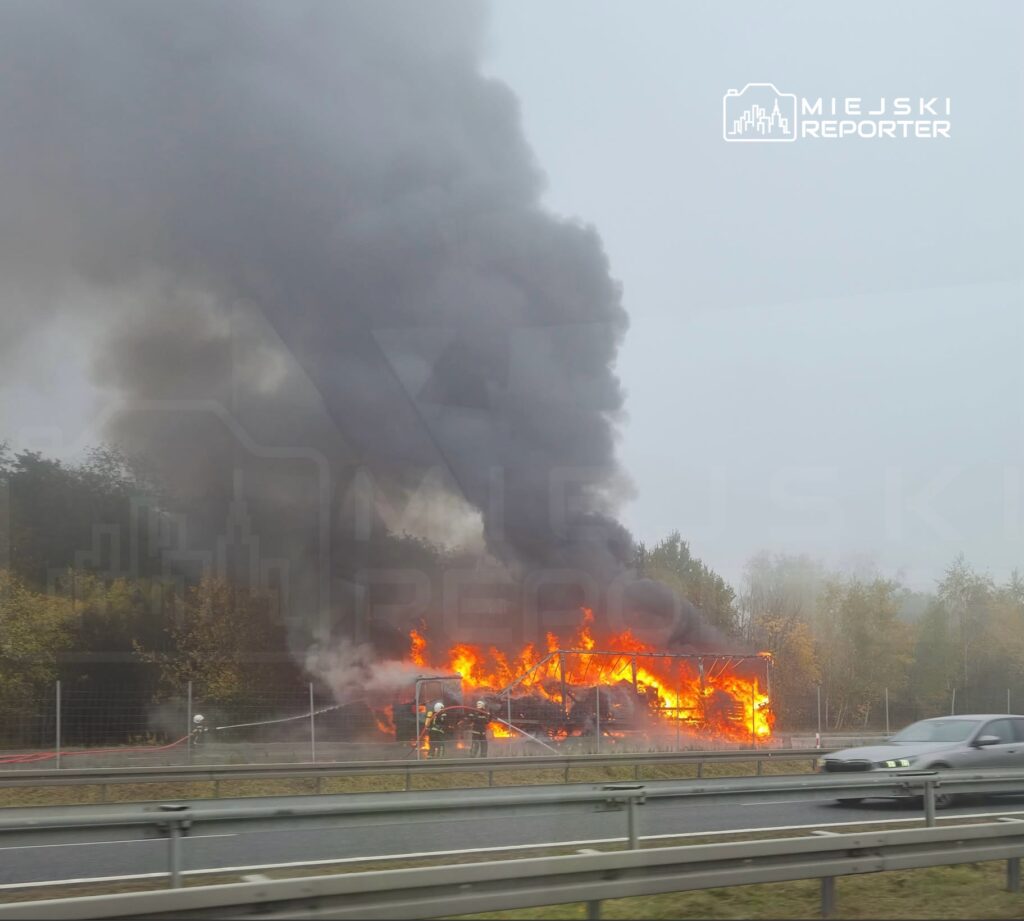 Płonący obiekt za ogrodzeniem przy drodze, nad ogniem unosi się gęsty, czarny dym.