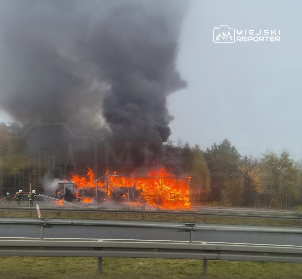 Strażacy gaszą płonący pojazd przy drodze; nad ogniem unosi się gęsty, czarny dym.