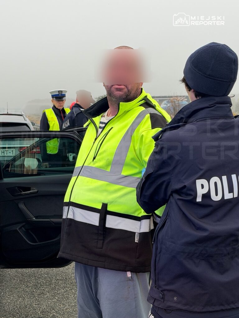 Policjant rozmawia z mężczyzną w kamizelce odblaskowej przy otwartych drzwiach samochodu, na parkingu.