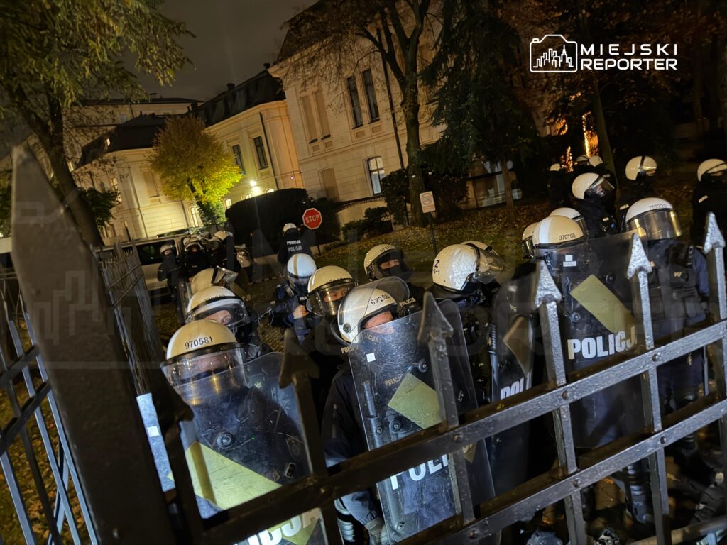 Funkcjonariusze policji w kaskach i z tarczami stoją za metalową barierką przed oświetlonym budynkiem nocą.