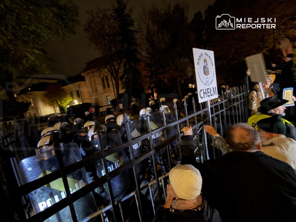 Policjanci w kaskach i z tarczami stoją za metalową barierą naprzeciw zgromadzonych osób; jedna trzyma tablicę „CHEŁM”.