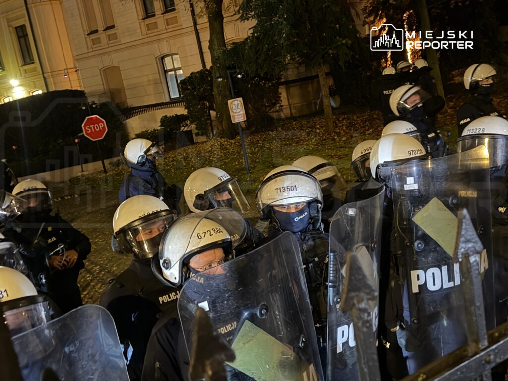 Umundurowani policjanci w kaskach i z tarczami stoją w szyku na ulicy przed oświetlonym budynkiem nocą.