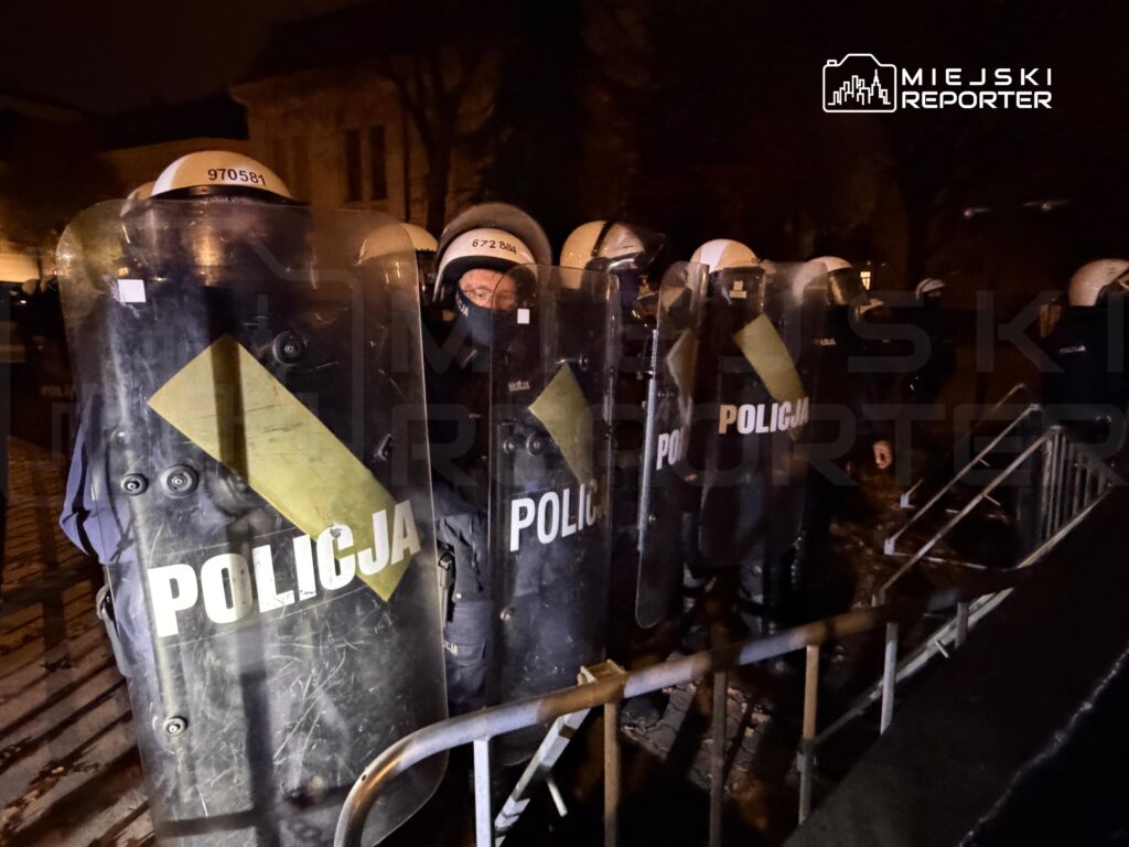 Funkcjonariusze policji w kaskach i z tarczami stoją nocą za metalową barierką na ulicy.