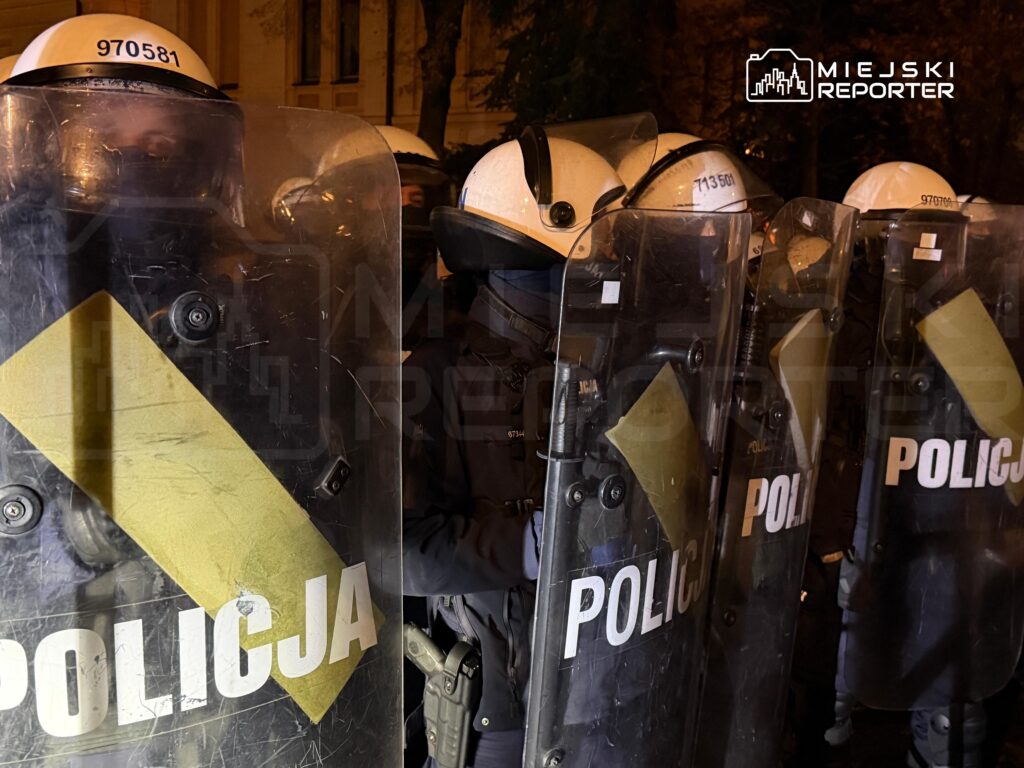 Policjanci w kaskach i z tarczami z napisem „POLICJA” stoją w szeregu nocą przy ulicy.