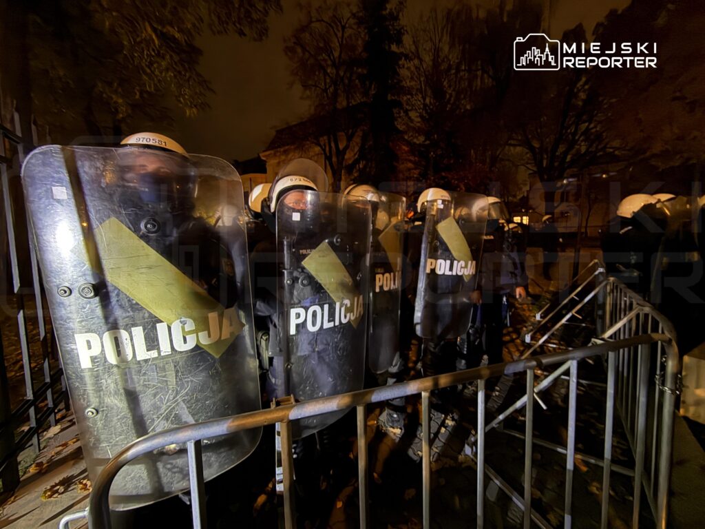 Funkcjonariusze policji w kaskach i z tarczami stoją za metalowymi barierkami nocą.